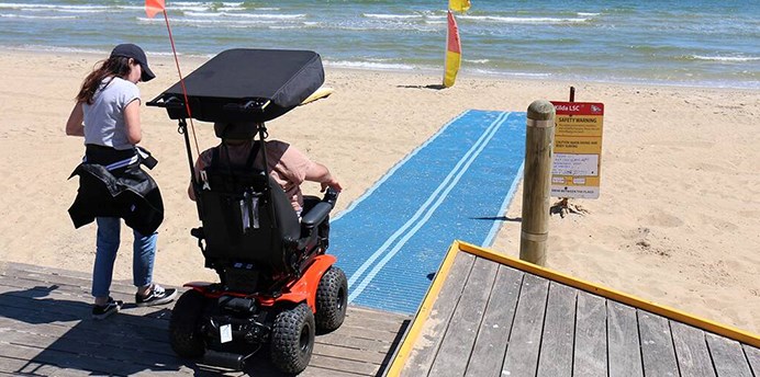 Person in a motorized wheel chair about to roll onto beach matting to access the water at St Kilda Beach