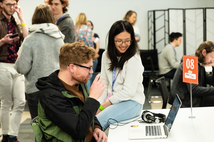 A group of young people male and female gathered some seated some standing talking and sharing computer screens looking at games.