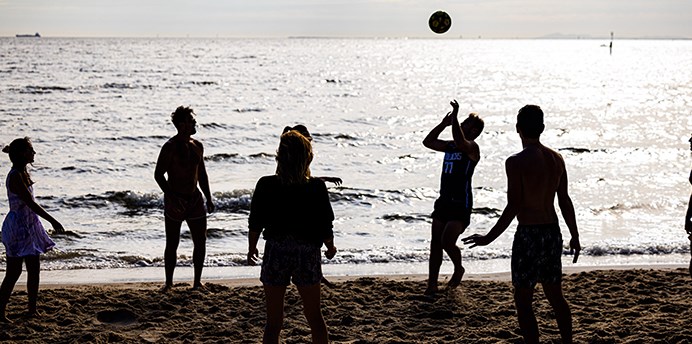 Group of 6 people in shadows play volleyball at St Kilda Beach while the sun sets behind them