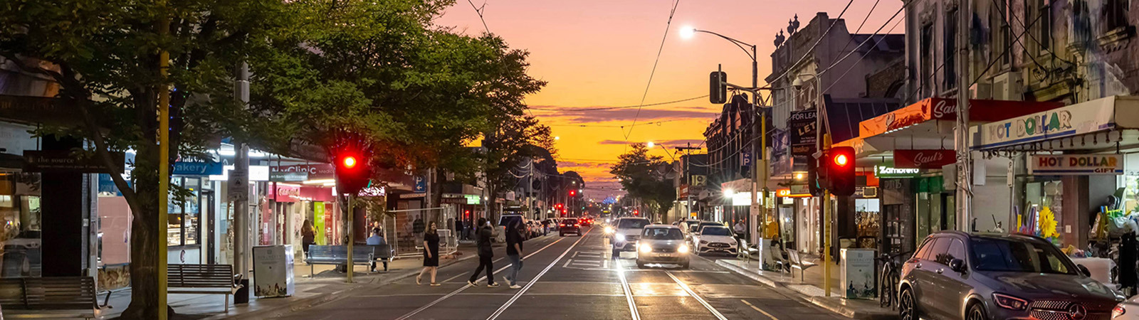 A view along Carlisle Street at sunset, looking down the centre of the road with tram tracks leading into the distance. Cars travel through the shopping strip while pedestrians cross at an intersection. Shopfronts, trees and streetlights line both sides of the street. Red traffic signals stand out against a warm orange and pink sky, with building lights beginning to glow at dusk.