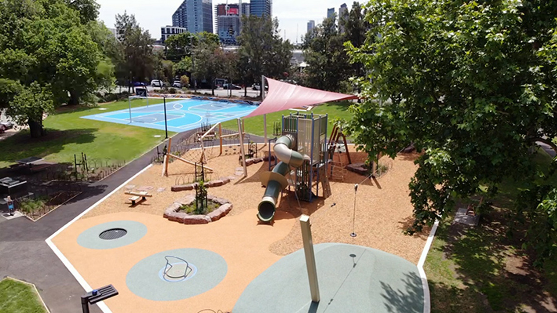 Elevated view of a playground with tan rubber flooring, climbing structures, slides, and a large pink shade sail. The playground is surrounded by trees and green grass, with a blue basketball court visible in the background. Tall buildings can be seen in the distance beyond the park.